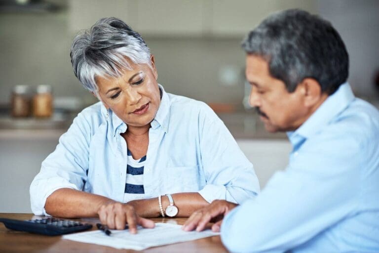 An older adult man and woman sit at a table, looking over paperwork. There is also a calculator and a pen on the table.