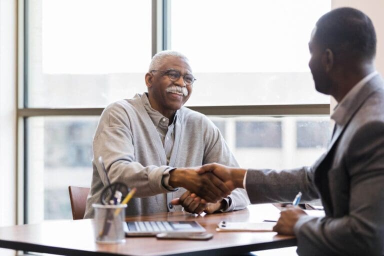 An older adult man shakes hands with a younger man. They both sit at a desk with a laptop and some paperwork between them.