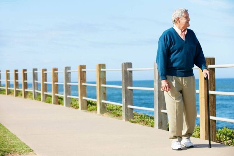 An older adult man walks along a path next to a body of water, using a cane.