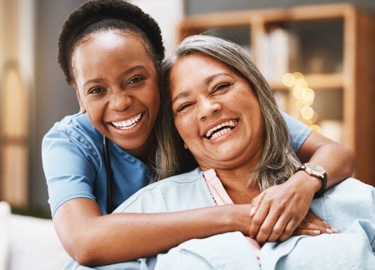 A woman health care provider stands beside a seated older adult woman, hugging her. They both smile at the camera.