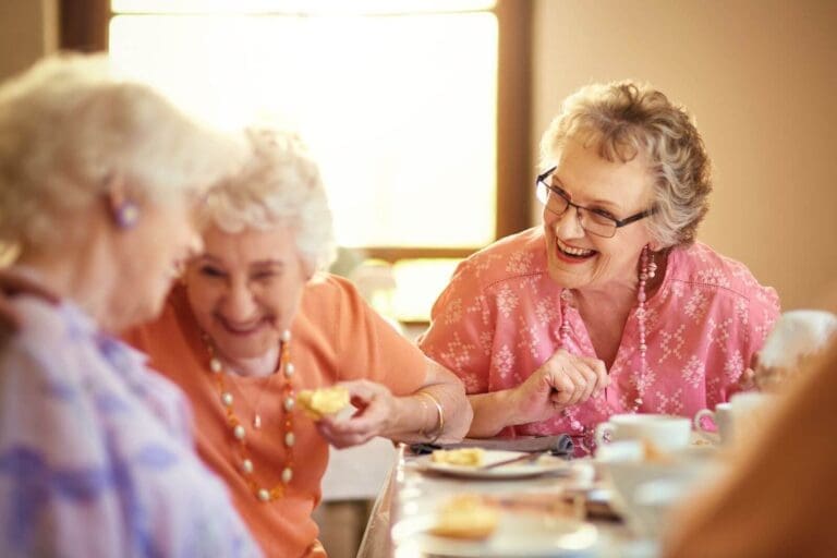 Three older adult women sit at a table, eating pastries and drinking tea. They are laughing with one another.