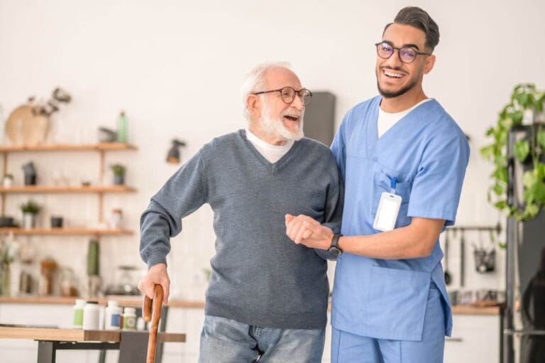 A man wearing scrubs helps an older adult man who is using a cane. He holds the older man's arm and smiles at the camera. The older man is smiling at the caregiver.