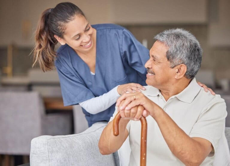 An older adult man holding a cane sits on a couch. He is turning to look at a younger woman caregiver, who has her hands on his shoulders.