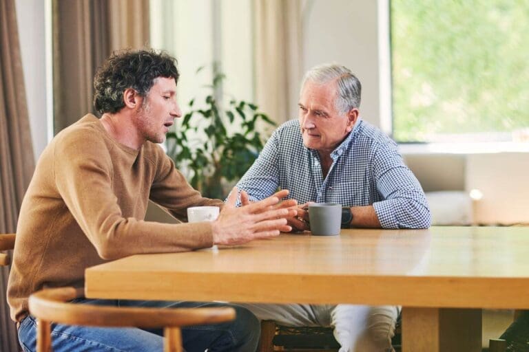 An older adult man and his adult son sit at a table talking. They both have coffee cups.