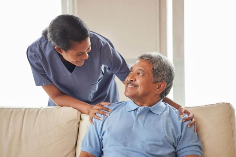 A female health care worker stands behind a man who is sitting on a couch. She has her hands on his shoulders, and they are smiling at each other.