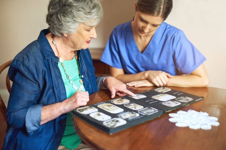 A woman caregiver and an older adult woman sit at a table looking at a book of old photographs.