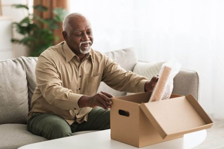 An older adult man sitting on a couch is placing a bubble-wrapped picture frame into a box on the table in front of him.