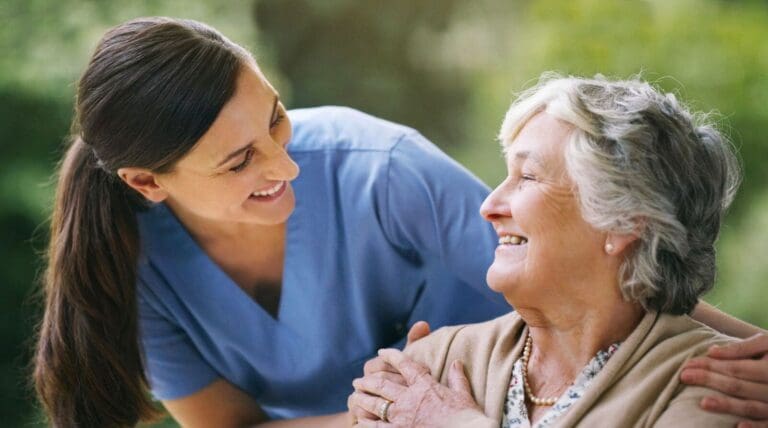 A female caregiver has her hands on the shoulders of a seated older adult woman. They smile at each other.