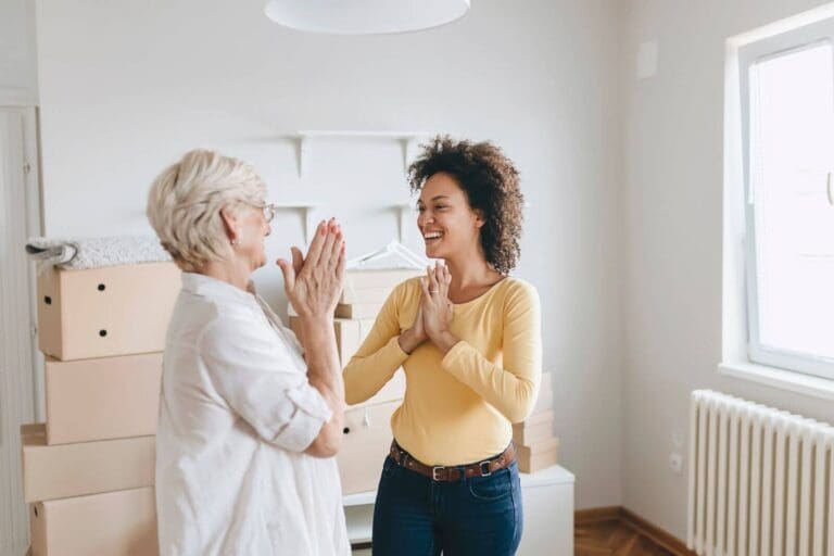 A woman and an older adult woman stand in a room full of cardboard boxes. They are laughing together.