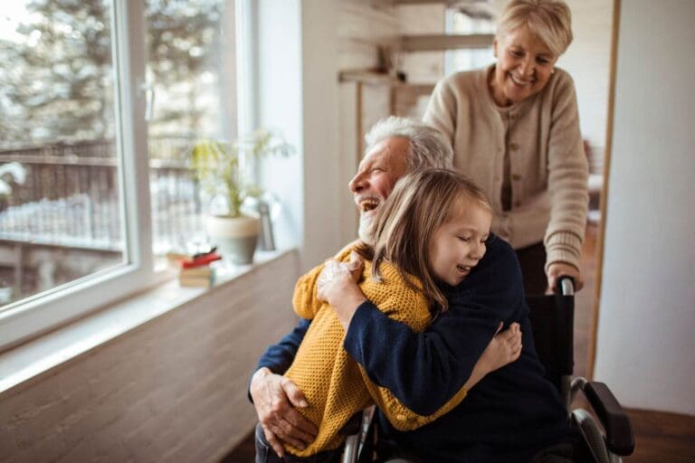 An older adult man sitting in a wheelchair hugs his granddaughter. An older adult woman is standing behind him, pushing the chair.