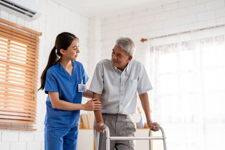 A female health care worker wearing scrubs assists an older adult man using a walker.