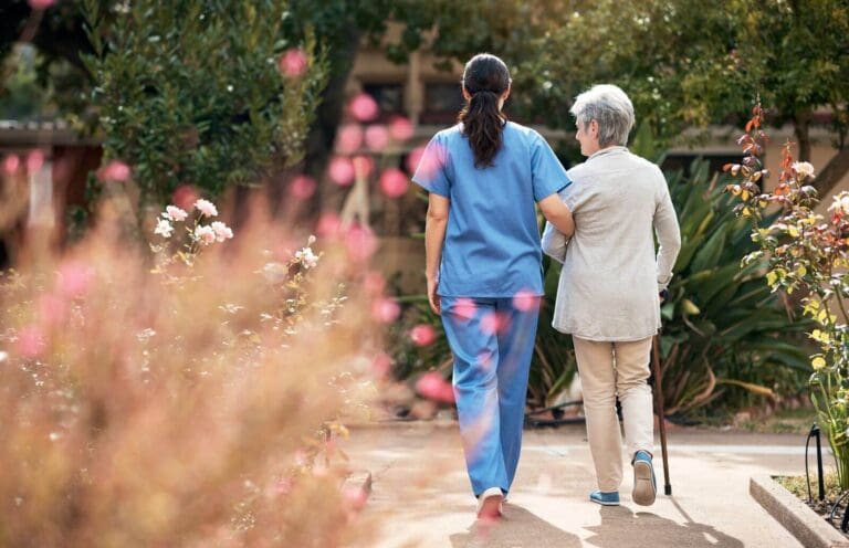 Shown from behind, an older adult woman walks outdoors with the help of a professional care aide.