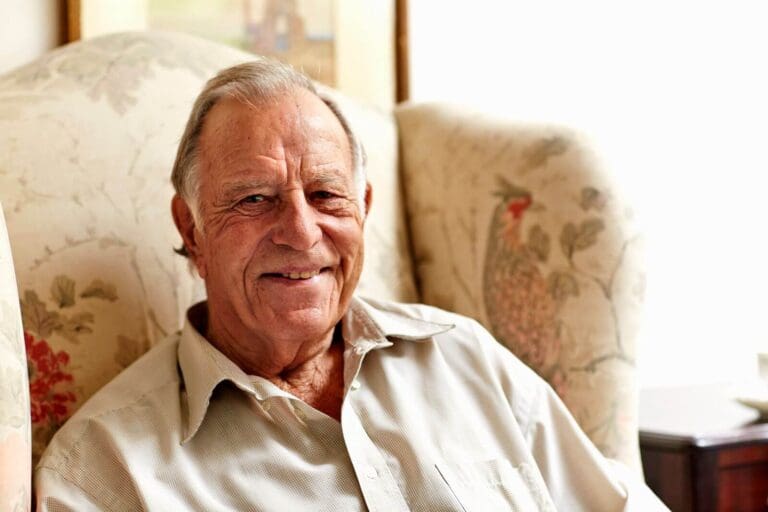 An older adult man sitting in an armchair smiles at the camera. He is in a sunny room.