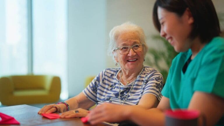 An older adult woman sits at a table with a female caregiver. They are folding origami.