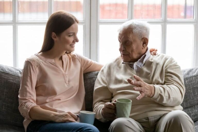 A woman sits on a couch talking to her older adult father. They are both holding coffee mugs, and she has her arm around his shoulders.