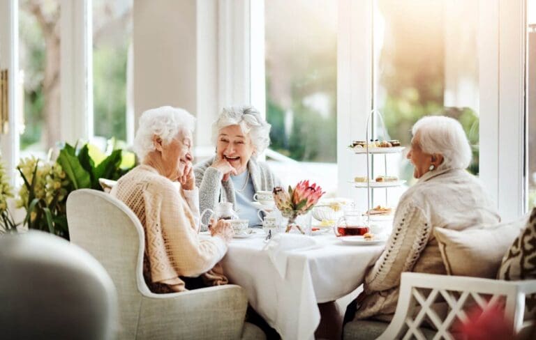 Three older adult women sit at a table drinking tea. They are laughing.