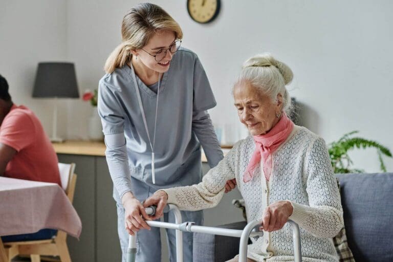 A female caretaker assists an older adult woman as she stands from a chair with the help of a walker.