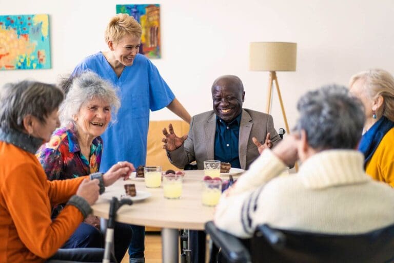 A group of five older adult men and women sit at a round table together with lemonade and cake in front of them. A female nurse stands with them, smiling.