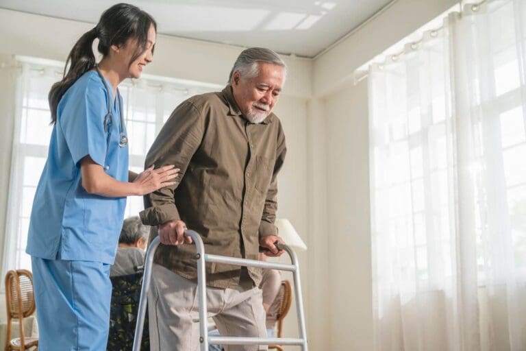 A female physical therapist helps an older adult man walk with the assistance of a walker.