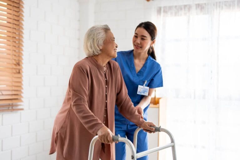 A female health care worker assists an older adult woman as she walks using a walker.