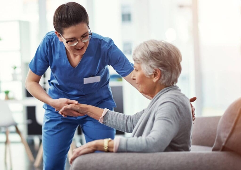 A woman aide wearing scrubs is helping an older adult woman to stand up from an armchair.