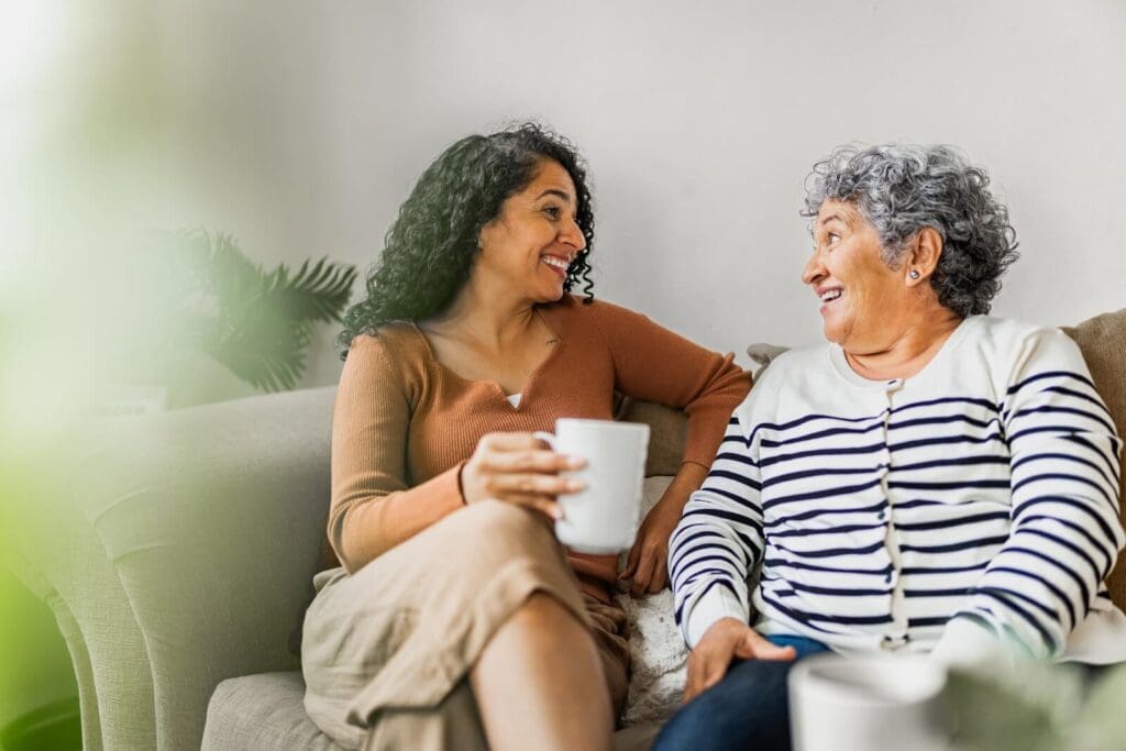 A woman is sitting on a couch, holding a coffee mug and talking with her older adult mother.