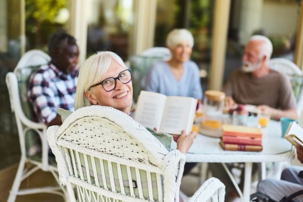 An older adult woman is seated at a table with four other older adults. She is holding a book and turned to look behind her, smiling at the camera.