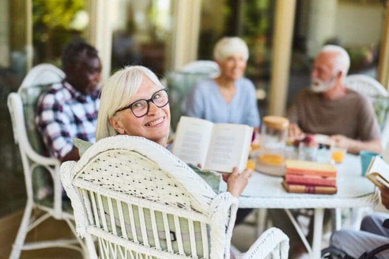 An older adult woman is seated at a table with four other older adults. She is holding a book and turned to look behind her, smiling at the camera.