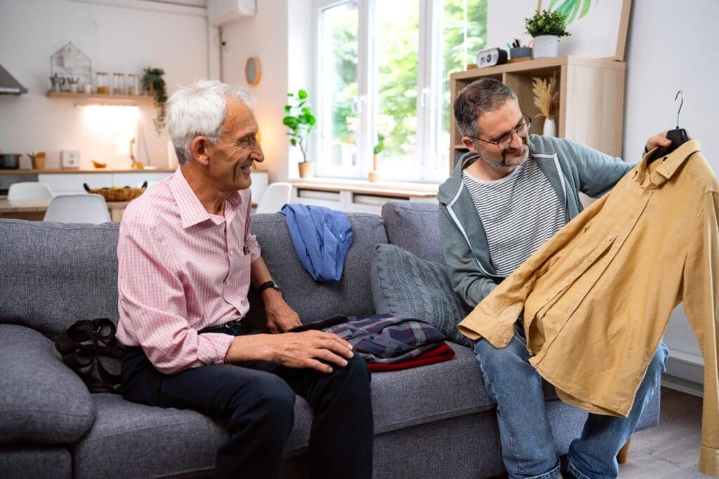 An older adult man and his son are sitting on a couch, packing clothes. The son holds up a button-down shirt to show his father. The older man is smiling.