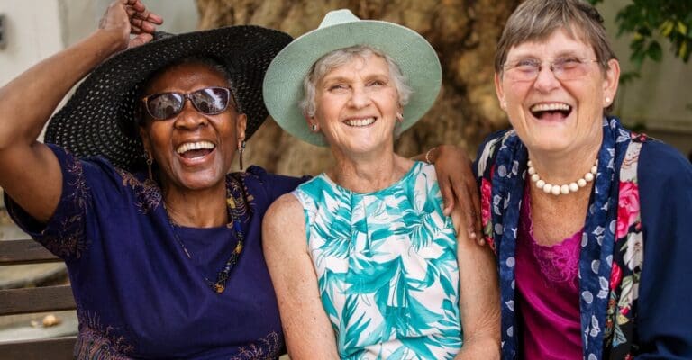 Three older adult women are sitting outdoors and smiling and laughing while looking at the camera.