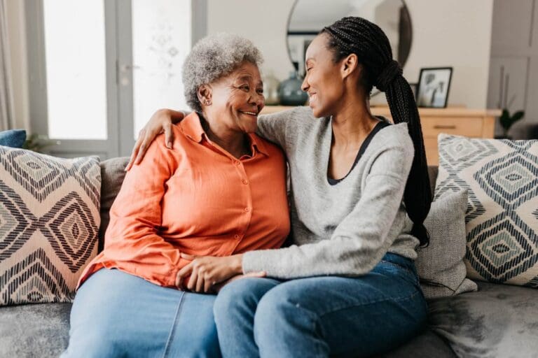 A woman sits on a couch with her older adult mother with her arm around her shoulders and holding her hand. They are smiling at each other.