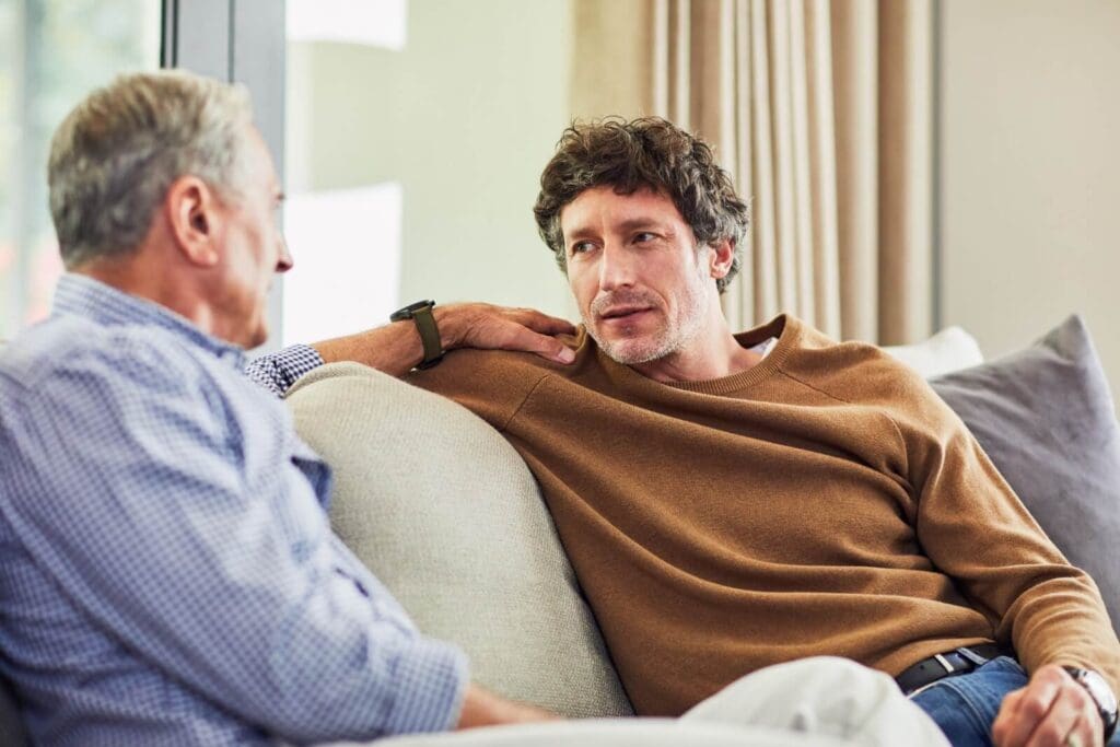 A man and his older adult father are sitting on a couch, having a discussion. The father has his hand on his son's shoulder.