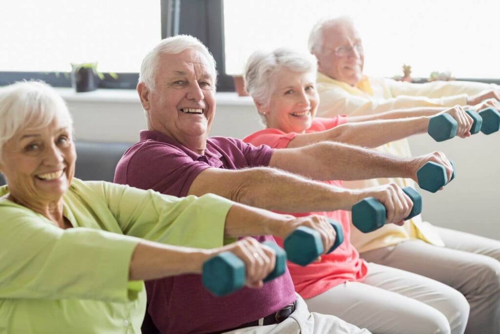 Two older adult men and two older adult women are seated, holding small dumbbells in each hand. They are looking at the camera and smiling.