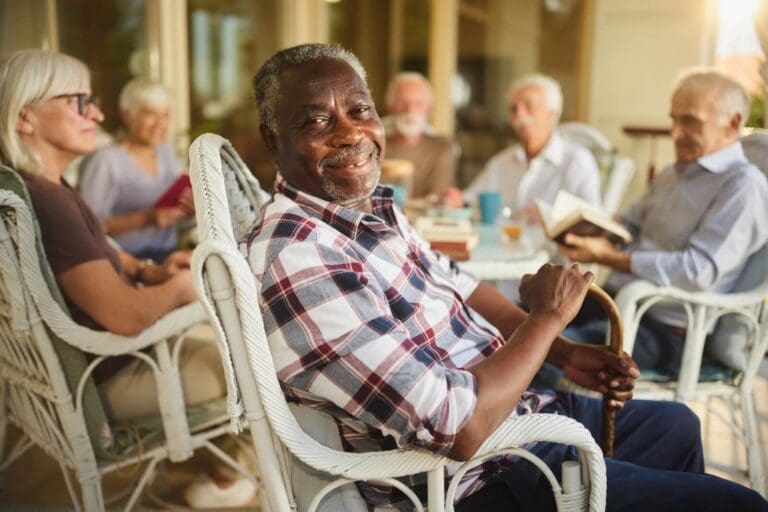 A group of four older adult men and two older adult women are sitting at an outdoor table. One man is turned around to smile at the camera. Some people are reading or chatting behind him.