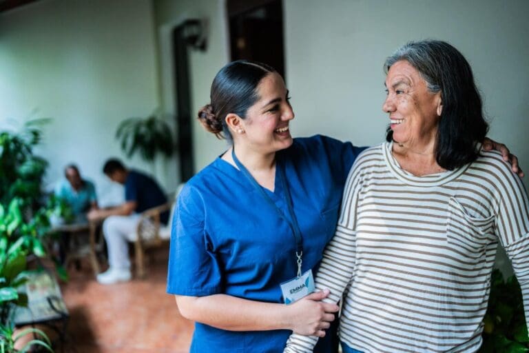 A woman caregiver holds the arm of an older adult woman and has her other arm around her shoulder. They are smiling at each other. There are two seated people in the background.