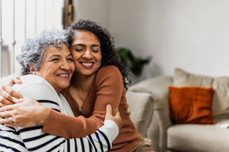A woman and her older adult mother sit on a couch, hugging each other and smiling.