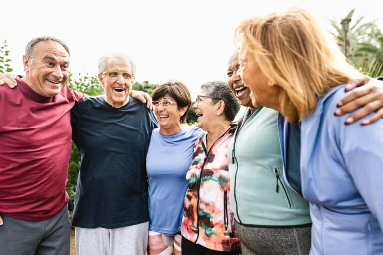 Two older adult men and four older adult women are standing with their arms around each other. They are all laughing.