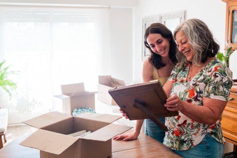 A woman and her older adult mother are standing in front of a table, looking at a framed picture the older woman is holding. There is a box on the table and two others in the background.