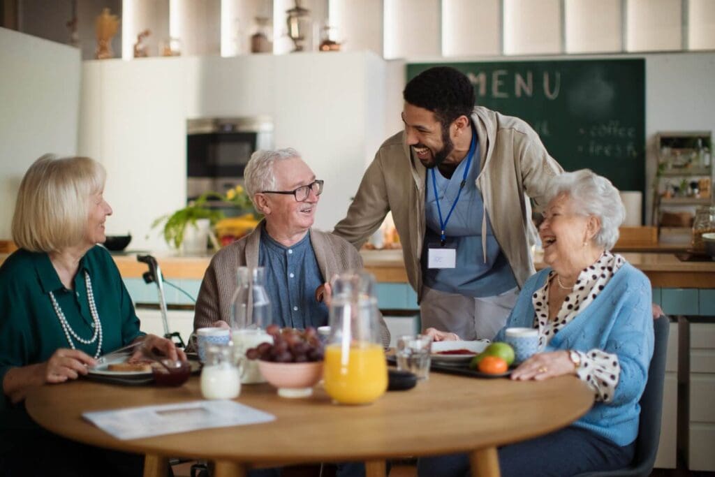 Three older adults sit at a table with plates of food in front of there. There is also a jug of orange juice on the table. A senior care facility staff member is leaning down to talk to them. Everyone is laughing.