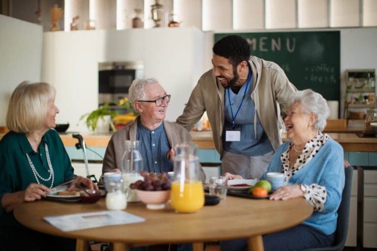 Three older adults sit at a table with plates of food in front of there. There is also a jug of orange juice on the table. A senior care facility staff member is leaning down to talk to them. Everyone is laughing.