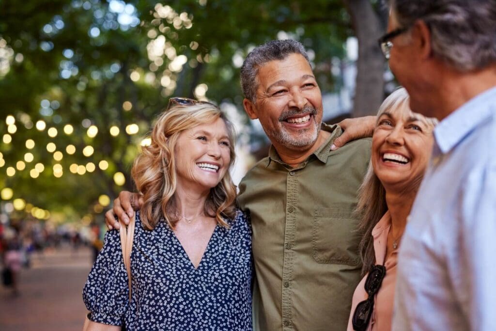 A group of two older adult men and two older adult women are standing outside together with their arms around one another, smiling.