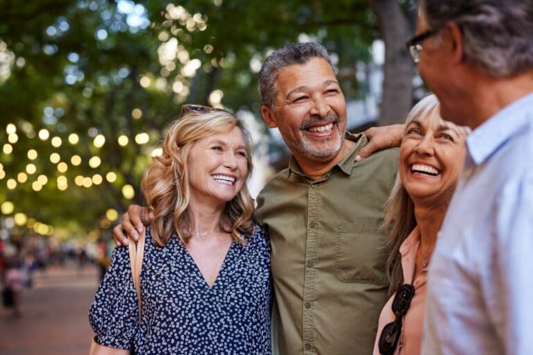 A group of two older adult men and two older adult women are standing outside together with their arms around one another, smiling.