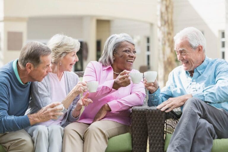 Two older adult men and two older adult women are sitting on patio furniture outdoors, drinking coffee and smiling at one another.