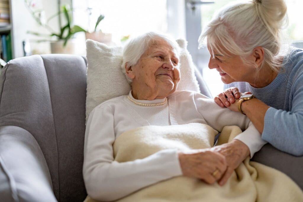 A woman leans down and smiles at her older adult mother, who is sitting in an armchair with a blanket on her lap.