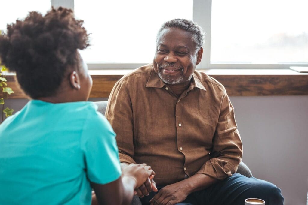 A woman sits with her older adult father, holding his hand. He smiles at her.