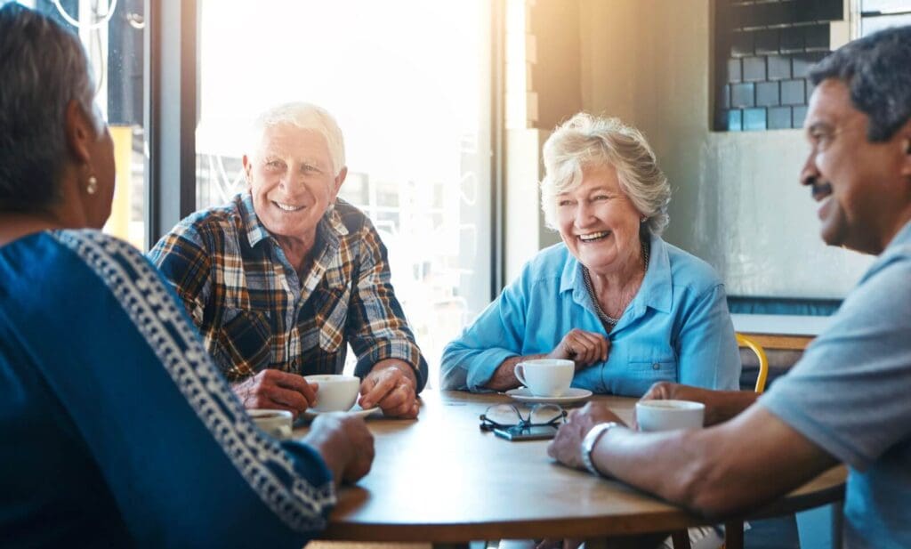 Two older adult women and two older adult men sit at a table, drinking coffee, talking, and smiling.