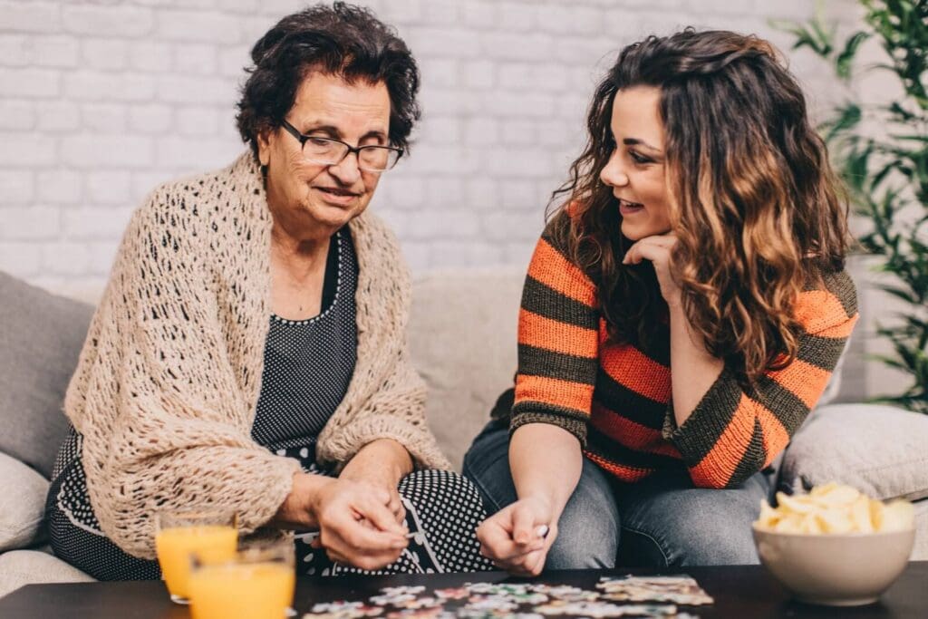 A young woman sits on a couch with an older adult woman. They are doing a puzzle together.