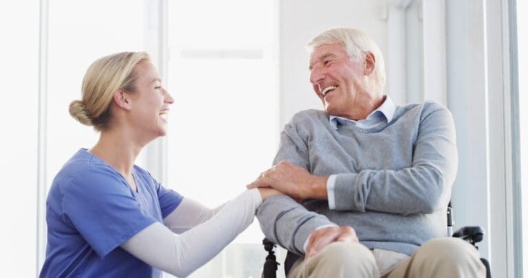 A female caregiver kneels next to an older adult man seated in a wheelchair. They are smiling at each other.
