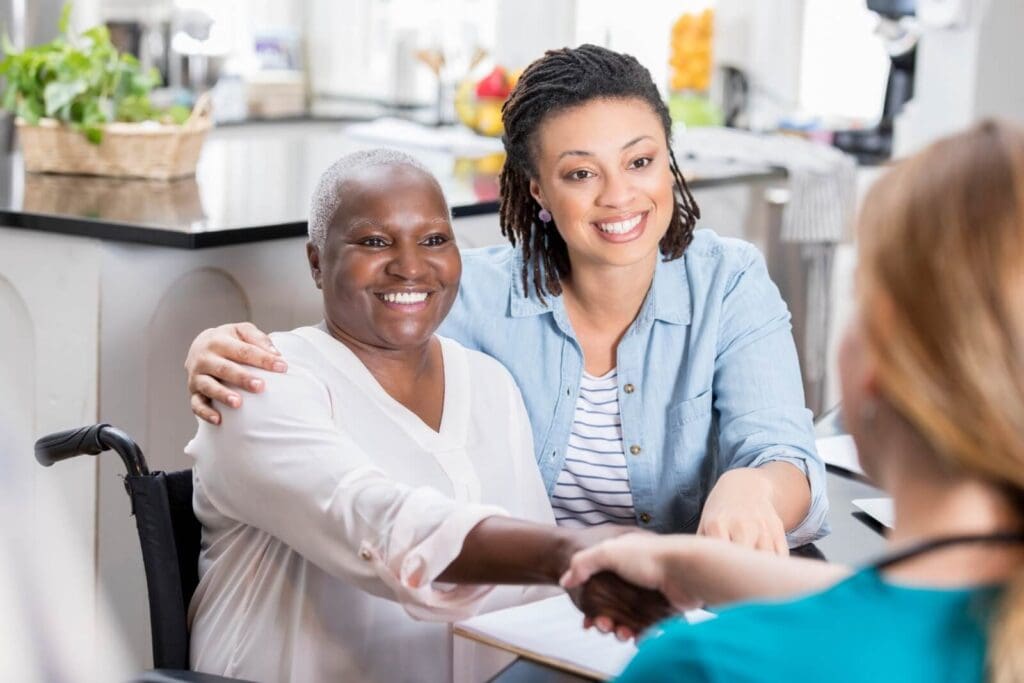 A woman, her older adult mother, and a health care worker sit at a desk. The older adult woman is seated in a wheelchair and shaking the hand of the health care worker.