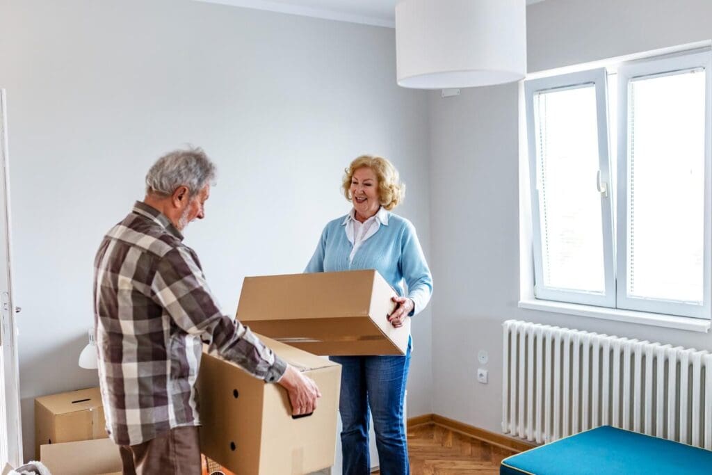 An older adult man and woman are standing, holding cardboard boxes. The room they are in has some other boxes but is quite empty, as they are moving to independent living.
